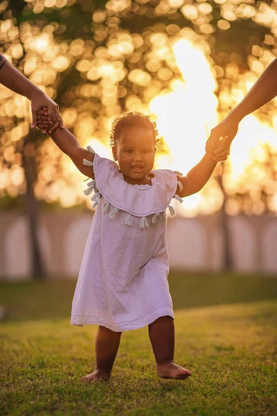 Positiva menina afro-americana de um ano e meio caminha ao redor do gramado e dá seus passos de mãos dadas de seus pais Imagem De Stock