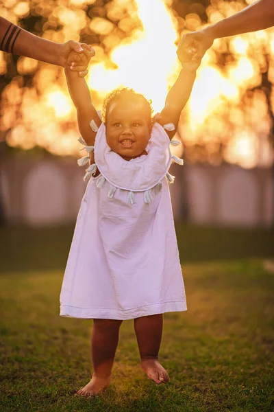 Positiva menina afro-americana de um ano e meio caminha ao redor do gramado e dá seus passos de mãos dadas de seus pais Imagens Royalty-Free