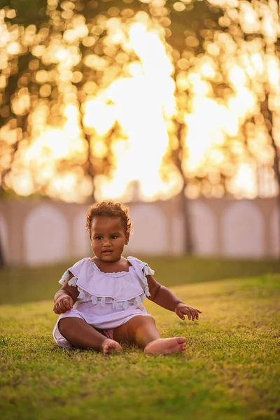 Doce menina afro-americana vestindo bonito vestido rosa e senta-se na grama ao ar livre Imagem De Stock