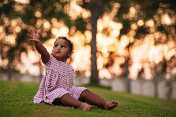 Doce menina afro-americana vestindo bonito vestido rosa e senta-se na grama ao ar livre com a mão para cima Imagem De Stock