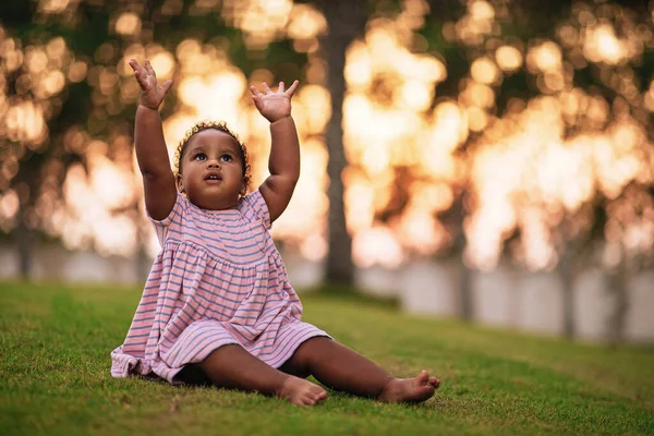 Doce menina afro-americana vestindo bonito vestido rosa e senta-se na grama ao ar livre com as mãos para cima Fotografia De Stock