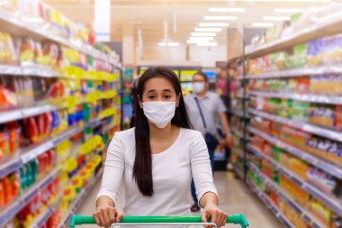 Asyalı bir kadın süpermarkette maske takıyor alışveriş arabasını itiyor. Bir şey almak için markete giden bir kız. Coronavirus krizi ya da kovid19 salgını sırasında. Koruyucu yüz maskesi takan kadınlar