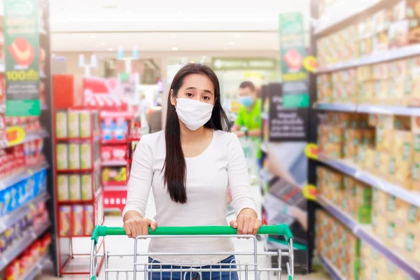 Asian woman wear face mask push shopping cart in supermarket department store. Girl looking grocery to buy  something. During coronavirus crisis or covid19 outbreak. Women wearing protective face mask