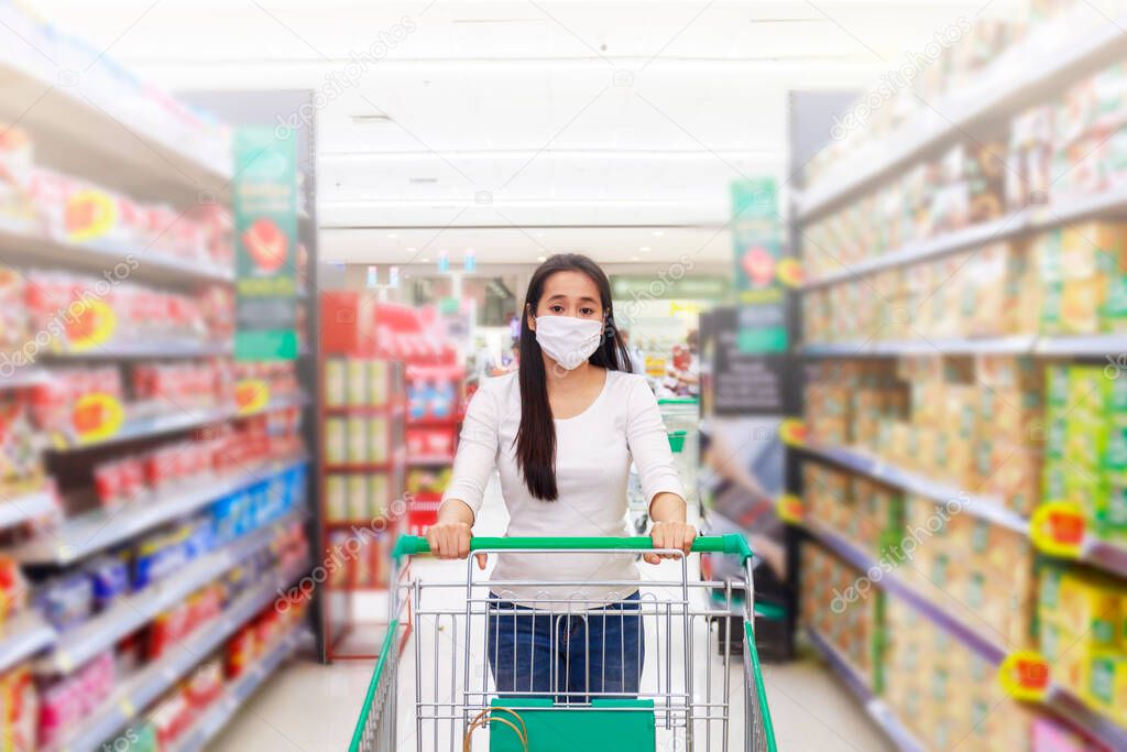 Asian woman wear face mask push shopping cart in supermarket department store. Girl looking grocery to buy  something. During coronavirus crisis or covid19 outbreak. Women wearing protective face mask