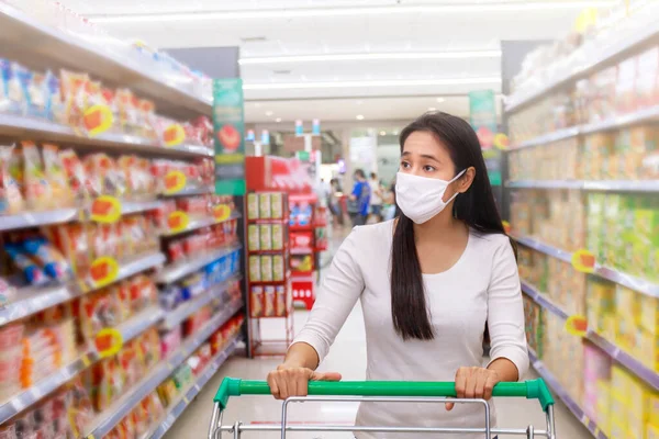 Asian woman wear face mask push shopping cart in supermarket department store. Girl looking grocery to buy  something. During coronavirus crisis or covid19 outbreak. Women wearing protective face mask