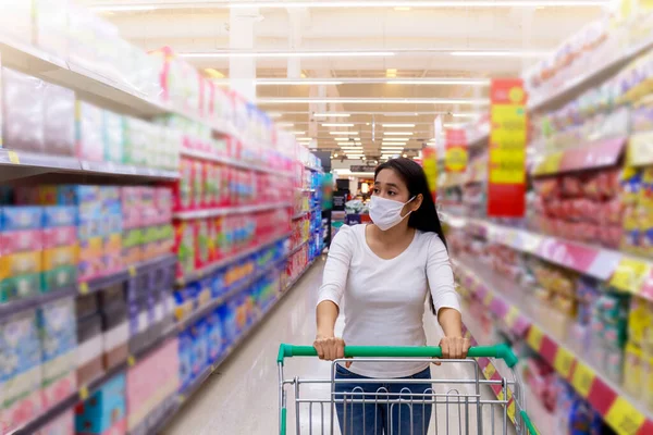 Asian woman wear face mask push shopping cart in supermarket. Girl looking grocery to buy  something. During coronavirus crisis or covid19 outbreak. Women wearing protective face mask new normal concept.