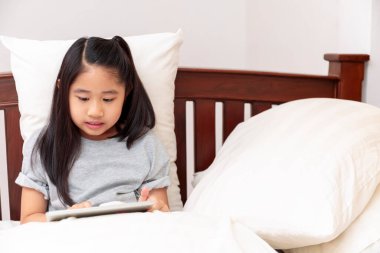 Yatak odasında el ele tutuşup tablet oynarken gülümseyen Asyalı küçük kız. Küçük kız yatakta tabletle eğleniyor. Asyalı kız arıyor ve tablet oynuyor..