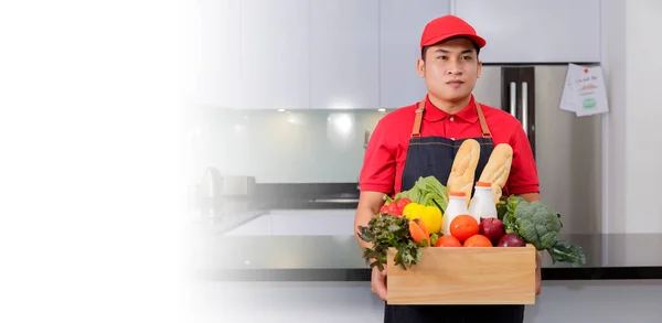 Grocery delivery courier man in red uniform and apron with grocery box with food, fresh fruit and vegetable. Deliver man handling wooden crate of food, fruit, vegetable with clipping path banner