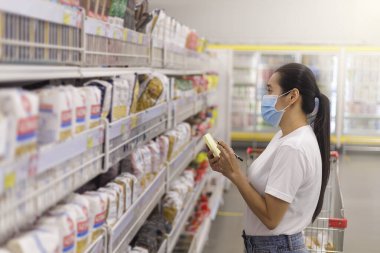 Asyalı kadın yüz maskesi takıyor süpermarkette alışveriş arabası itiyor. Coronavirus krizi ya da kovid19 salgını sırasında bir şeyler almak isteyen bir kız. Kadınlar yeni normal konsept listesindeki alışveriş malzemelerini kontrol ediyorlar