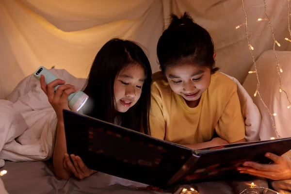 Family concept. elder sister and sister reading book with flashlight together in children tent before bedtime. Happy elder sister read story book to her sister in bed sheet tent — Stock Image Family Concept Elder Sister Sister Reading Book Flashlight Together Children Royalty Free Stock Photos
