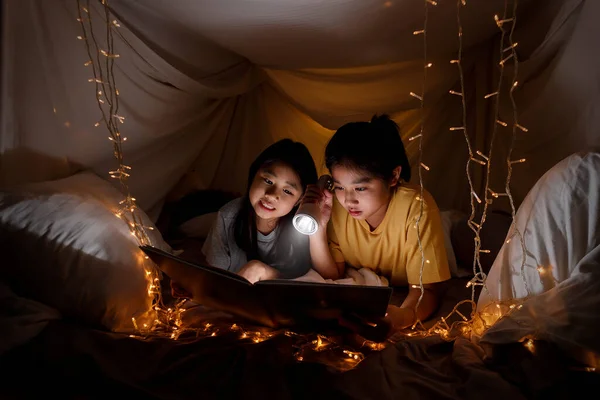 Family concept. elder sister and sister reading book with flashlight together before bedtime. Sister read story book together in bed sheet tent. focus selective little sister. With film grain effect — Stock Image Family Concept Elder Sister Sister Reading Book Flashlight Together Bedtime Royalty Free Stock Images