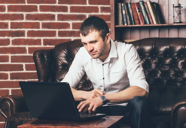 ハンサムな男は 本と棚の上の背景にノート パソコンの情報を読み取ります 実業家は彼の家の柔らかいソファの上に座っています — ストック写真