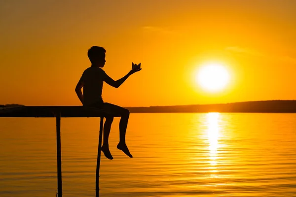 川の橋の上で夕日を背景に紙の船を持った少年のシルエットが描かれています — ストック写真