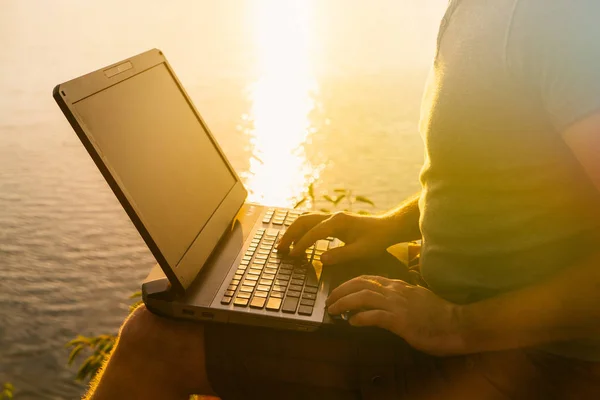 hands of man are typing a message on the laptop near the river at sunset. The rays of the evening sun fall on the laptop of man