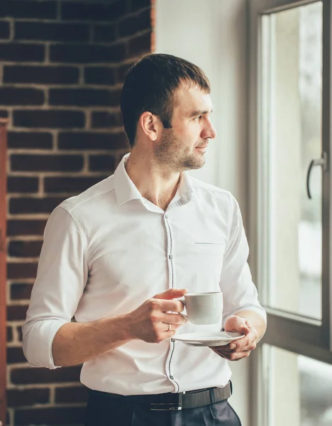 ビジネスマンは オフィスから彼の手でコーヒーのカップを持つウィンドウに見えます 外の出来事を観察します — ストック写真