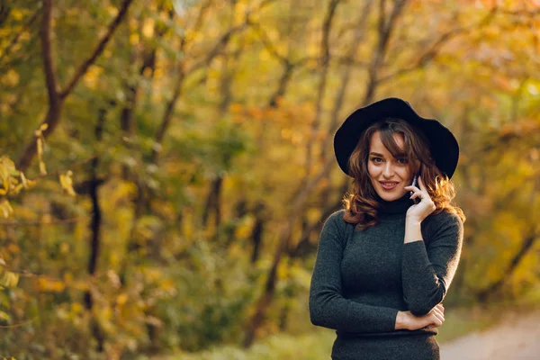黒い帽子の美しい少女は 秋に公園で彼女の手に携帯電話を保持しています 女性道路沿いの森の中を歩く — ストック写真