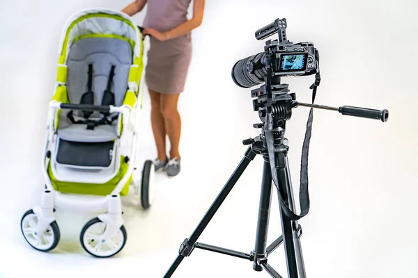 professional camera stands on a tripod and focuses on a woman in pink dress who holds stroller by her hands in the studio