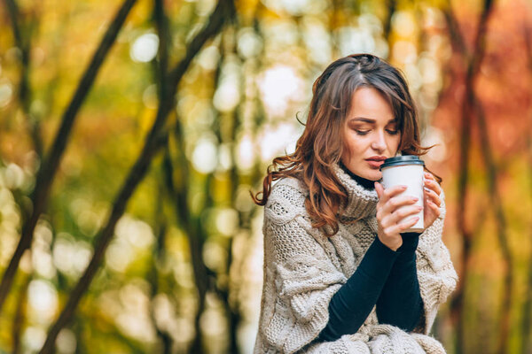 thoughtful woman in a knitted sweater holds a cup of hot coffee in the park in autumn