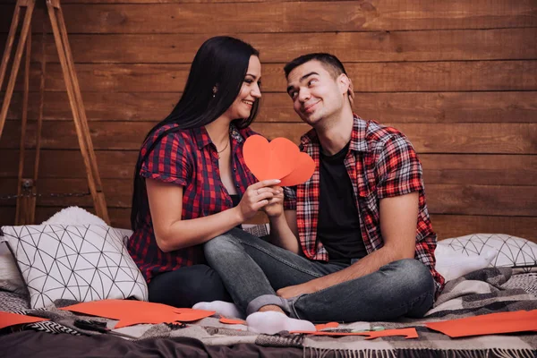 couple in love sits on the bed and red paper hearts on the Valentine\'s Day. Enamored man and woman in checkered shirts and jeans look at each other and flirt in the studio