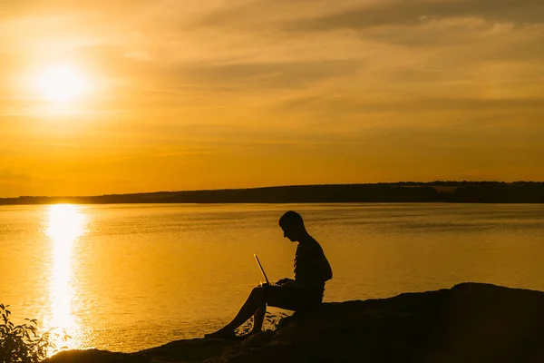 成人男性は 夏の夕日の背景に川の近くのラップトップで動作します 素晴らしい夜の風景 — ストック写真
