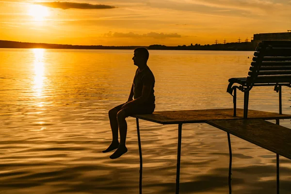 成人男性は 夏の夕暮れ時川のバック グラウンドのベンチで 石積みに座っています 素晴らしい景色 — ストック写真