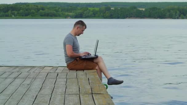 坐在老码头上的年轻人在笔记本电脑上工作 — 图库视频影像