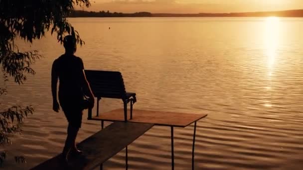 川沿いに木製の桟橋の上を歩く男のシルエット 地平線に息をのむの黄金の夕日 — ストック動画