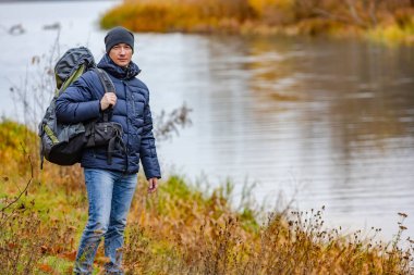 Sırt çantalı genç bir adam nehrin yanında duruyor. Yürüyüş için güzel sıcak sonbahar havası.