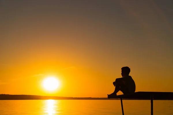 少年の空の夕焼けで川の橋の上に座ってのシルエット 孤独な少年が静かに座っているし 夕日の美しい景色の先に見える — ストック写真