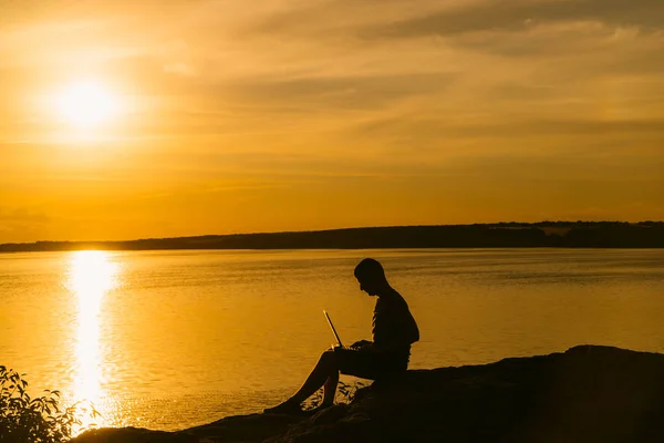 深刻な男は 夕暮れ時の彼の休暇の間に彼のラップトップで動作します 美しい夜の風景の背景以上のラップトップで男性の作業のシルエット — ストック写真