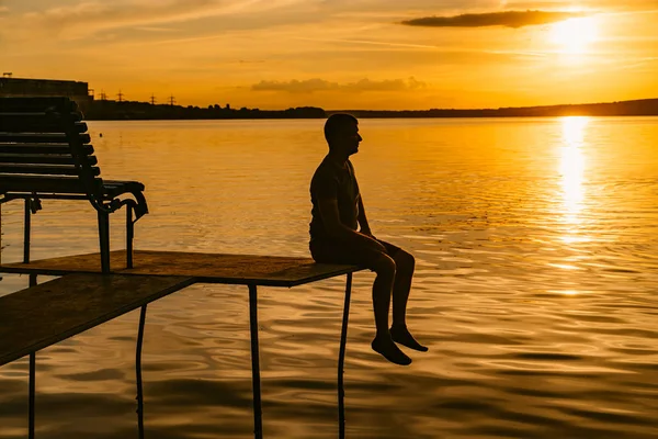 日没で木製の橋の上に座って一人で男のシルエット 小さな橋の上の孤独な男を見て明るい夕日太陽水の反映 — ストック写真