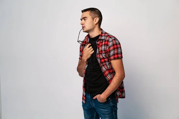Young male with eyeglasses to lips thinking a deep thought on white background. A portrait of handsome man in shirt and jeans with glasses in his hand and mouth. Concept problem solving.