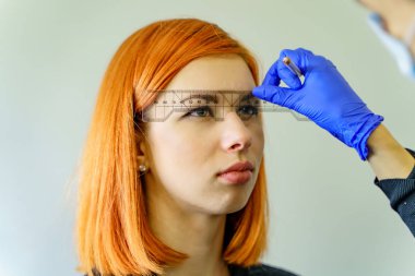 Stylist measuring the eyebrows with the ruler on a red-haired woman over the white background. Micropigmentation work flow in a beauty salon. clipart