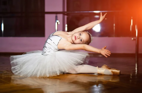 迷人的小女孩芭蕾舞演员在白色 Tutu 表演芭蕾姿势在地板上的工作室背景 一个可爱的小芭蕾舞演员坐在舞池里的舞蹈工作室里 — 图库照片