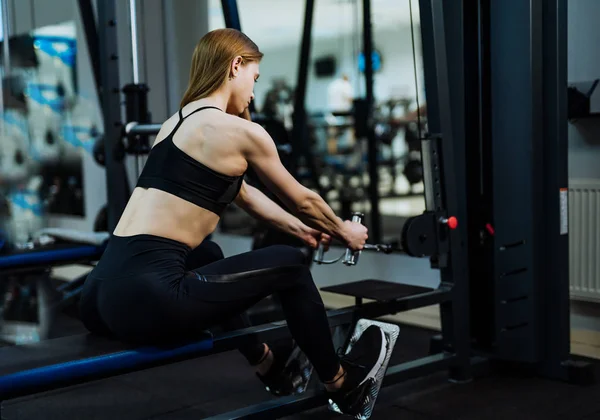 身穿黑色运动服和运动鞋的肌肉发达的年轻女子正在做密集的锻炼 在健身房拉着力量 运动女性正在体育俱乐部的模拟器上进行肌肉训练 — 图库照片