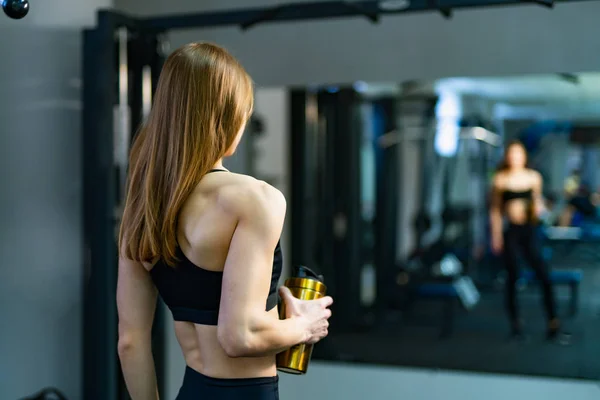 Beautiful athletic girl dressed in black sports top and tights wth shaker looks at her muscles in front of the mirror. Woman looking at the mirror after workout in a gym.