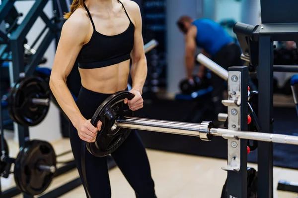 Professional sport fitness woman putting weight disk to barbell in gym. Athletic girl is preparing to workout with a barbell.