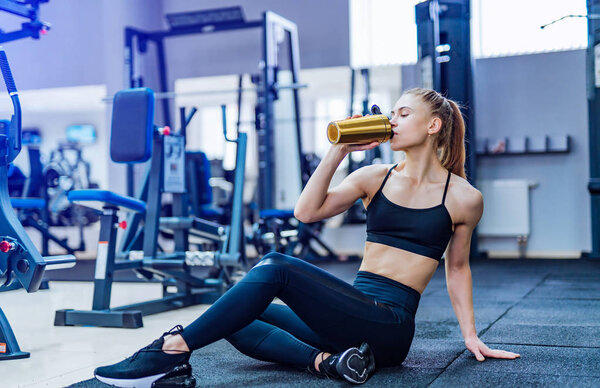 Fitness woman drinking water while sitting and resting on the floor in gym. Woman exercise workout in fitness gym breaking relax and holding protein shake bottle during training.