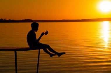 Tek başına bir ahşap duvar üzerinde oturmak ve nehrin yanında kağıt teknesinde batımını küçük erkek silüeti. Gün batımında köprüdeki origami ile çıplak çocuk.