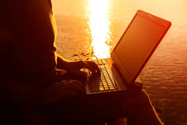 Businessman typing modern laptop against beautiful sunset landscape in the evening. Silhouette of a male sits near the river and works on a laptop. Close-up