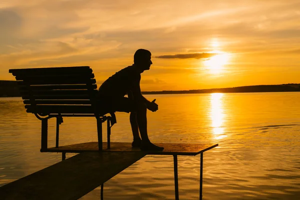 夕方には小さな橋のベンチに座って 夕日を見ている一人の男 座っていると 一人で川 湖または海で木製のベンチで休んで人間観 — ストック写真