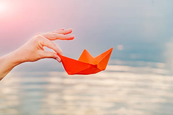 Hand of a young woman holding a paper boat by her fingers above the river in the summer. Orange paper boat in woman\'s hand. Close-up