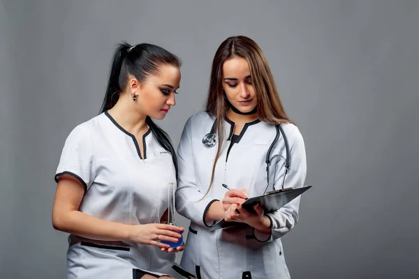 Two lovely doctors in medical uniform looking at the folder and discussing diagnosis on grey background. Nice women in medical clothes with black folder and test glass.