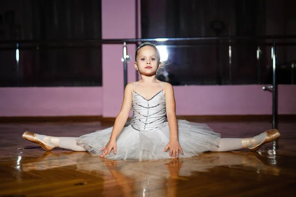 Lovely little ballerina in white tutu doing twine isolated on studio background. Portrait of flexible ballerina sitting on the floor and showing the twine