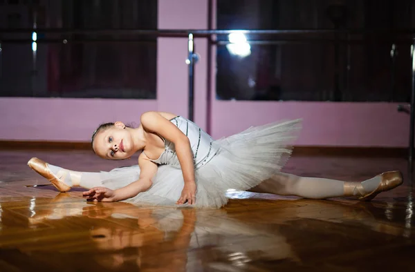 穿着白色西装的漂亮的小芭蕾舞演员在舞蹈工作室里做绳子 灵活的芭蕾舞演员在木地板上显示绳子 弯曲到一条腿 — 图库照片