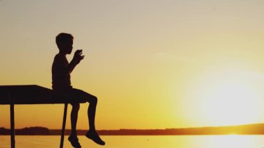 Çocuk duvar ucunda oturan ve gün batımında çakıl suya atma