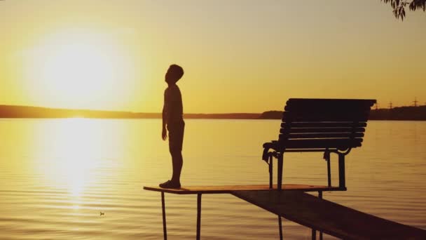 在河边日落的背景下 男孩正在板凳附近的桥上做体育活动 — 图库视频影像