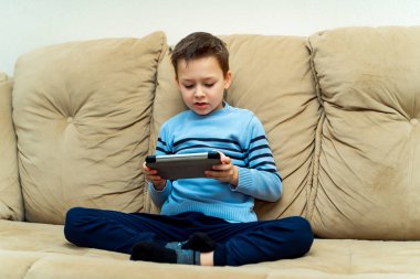 Rahat bir kanepede oturan ve bir tablet üzerinde oyun rahat kıyafetler hevesli küçük çocuk. Dijital tablet kanepede onun elinde çocukla