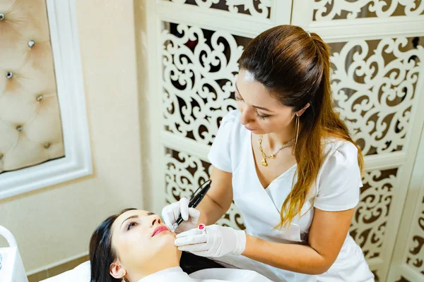 Pretty cosmetologist applying permanent make up on girl lips in beauty studio. A young woman having red lips permanent makeup.