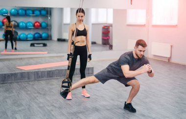 Trx ve spor salonunda gözü üstünde bir kadın ile egzersiz yaparak güçlü sakallı eğitmeni. Çok kadın ayakta bacaklar Trx. spor konsepti üzerine eğitim yapan yakışıklı erkek yakınındaki